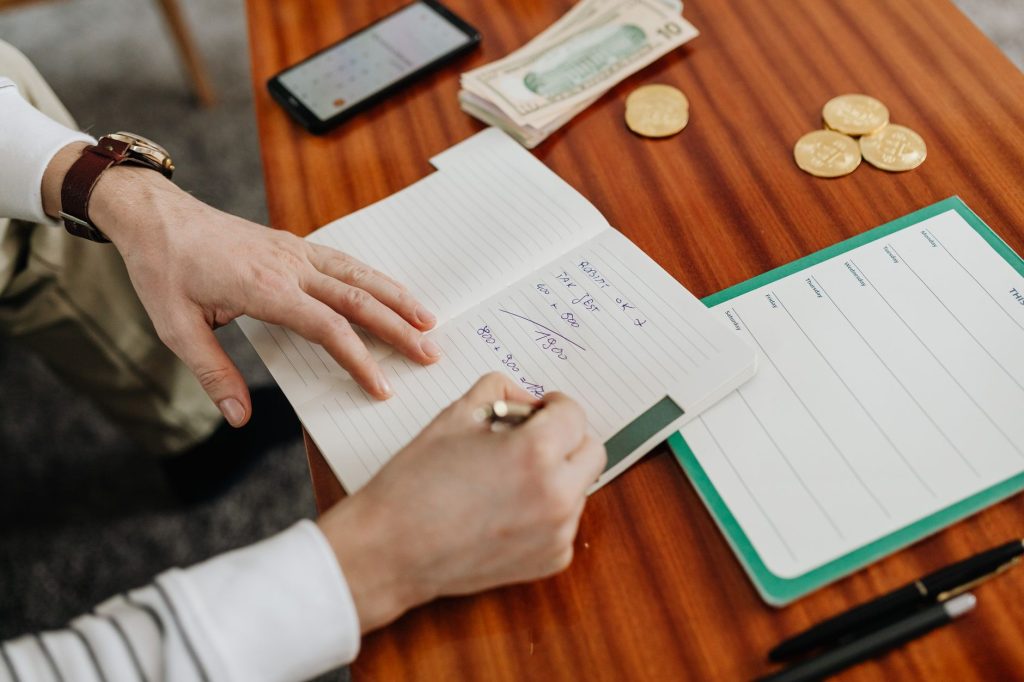 Mãos fazendo cálculos em um caderno ao lado de cédulas e moedas sobre uma mesa.
