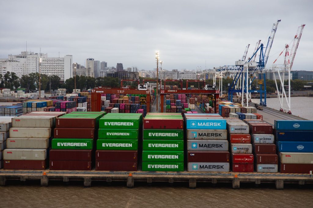 Acordo Mercosul-EFTA entra em vigor e amplia oportunidades comerciais para empresas brasileiras Vista aérea de contêineres coloridos empilhados em um movimentado porto, com guindastes e embarcações ao fundo.