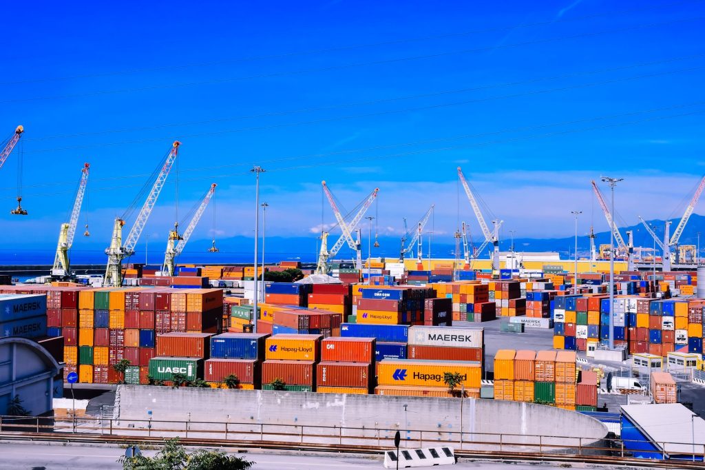 Porto industrial com contêineres empilhados e guindastes de carga sob céu azul.