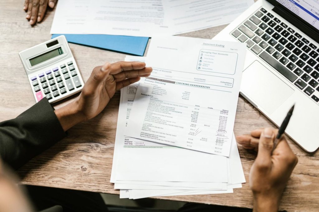 Mãos analisam documentos financeiros ao lado de uma calculadora e um laptop em uma mesa de escritório.